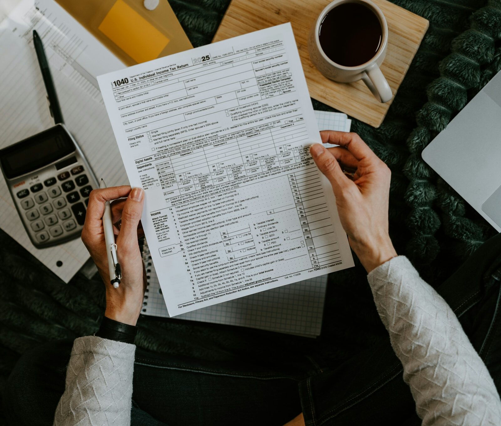 A person filling out IRS Form 1040 at a desk with a calculator and laptop